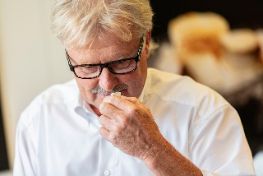 Das Bild zeigt den ehemaligen Geschäftsführer Jörg Baumann, einer der Söhne des Gründers. Er hat blondes Haar und trägt eine schwarze Brille. Er lächelt und riecht an einem Stück Käse.